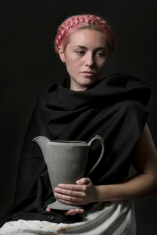 Young woman with braided dyed pink hair posing for a portrait holding a gray pitcher